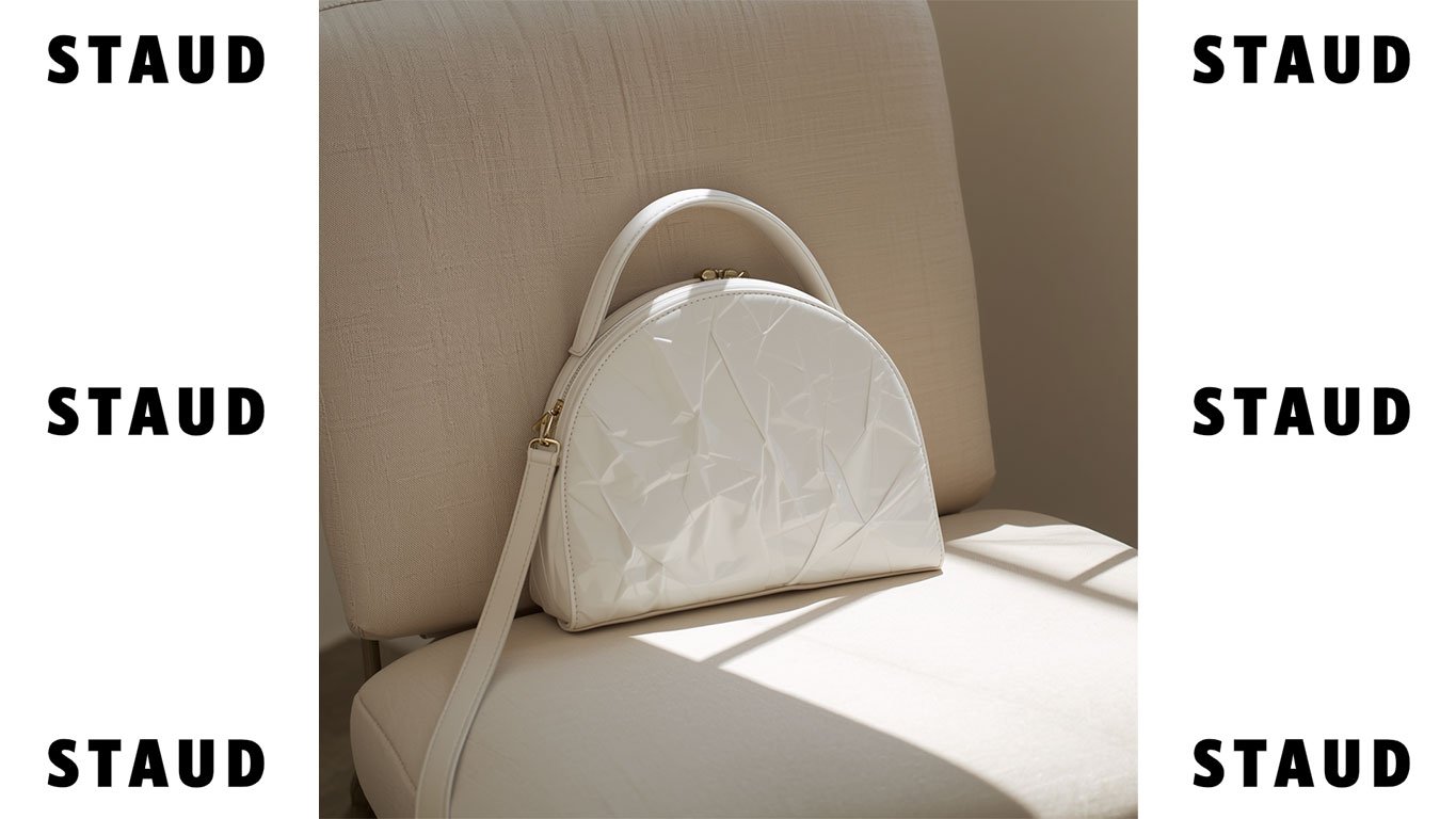 Staud white crinkled patent leather bag. Half-moon luxury handbag with top handle and gold zipper details on a minimalist background. STAUD logo branding on the left and right margins.