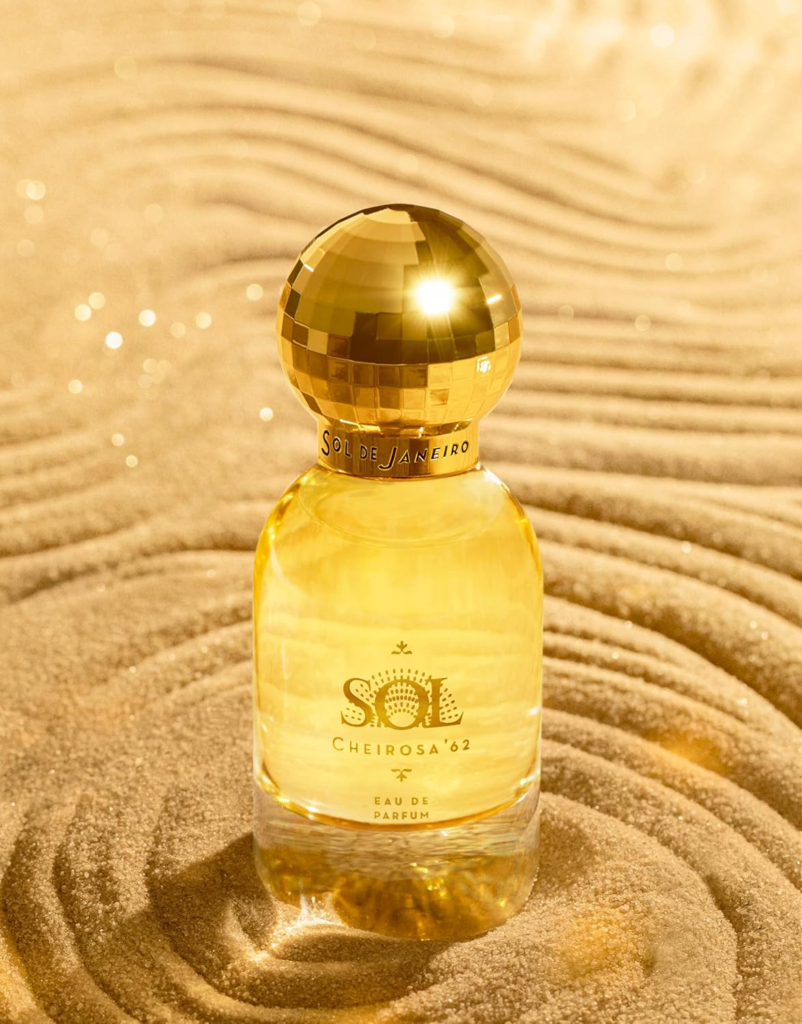 The iconic yellow bottle of Sol de Janeiro Cheirosa 62 Perfume Mist, featuring a white textured cap. The backdrop includes tropical elements like a coconut and warm sand, reflecting its pistachio and salted caramel scent profile.